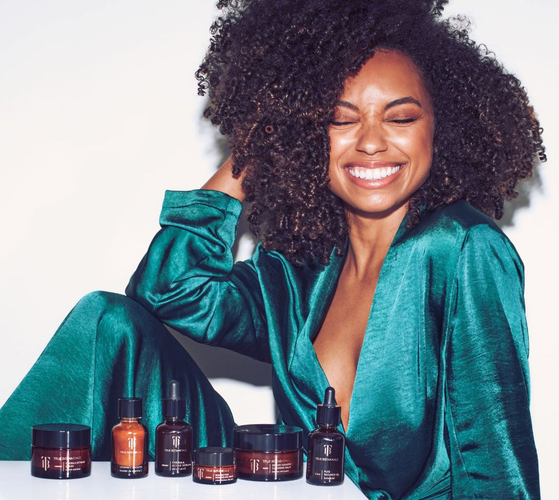 Smiling woman in a vibrant green satin outfit sits beside a full lineup of True Botanicals skincare products, including serums, creams, and oils, displayed in amber jars and bottles against a white background.