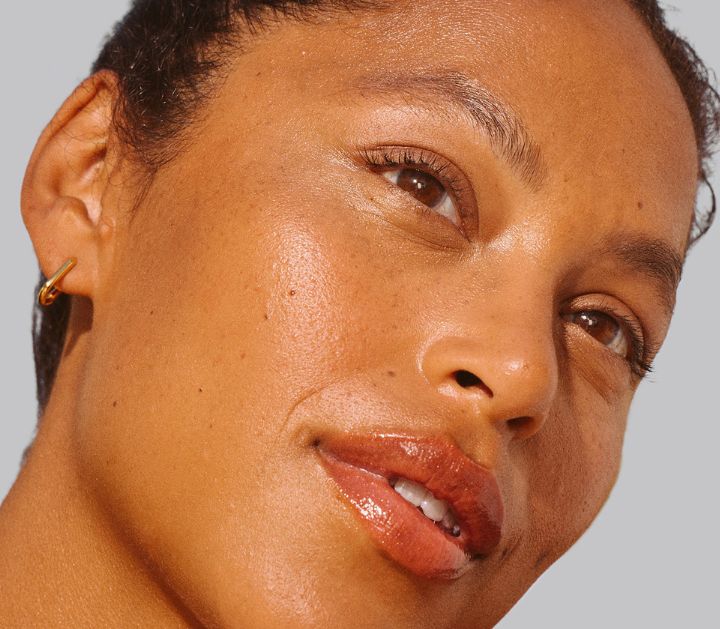 Close-up of a woman with glowing skin, warm brown eyes, and glossy lips, wearing a gold hoop earring.