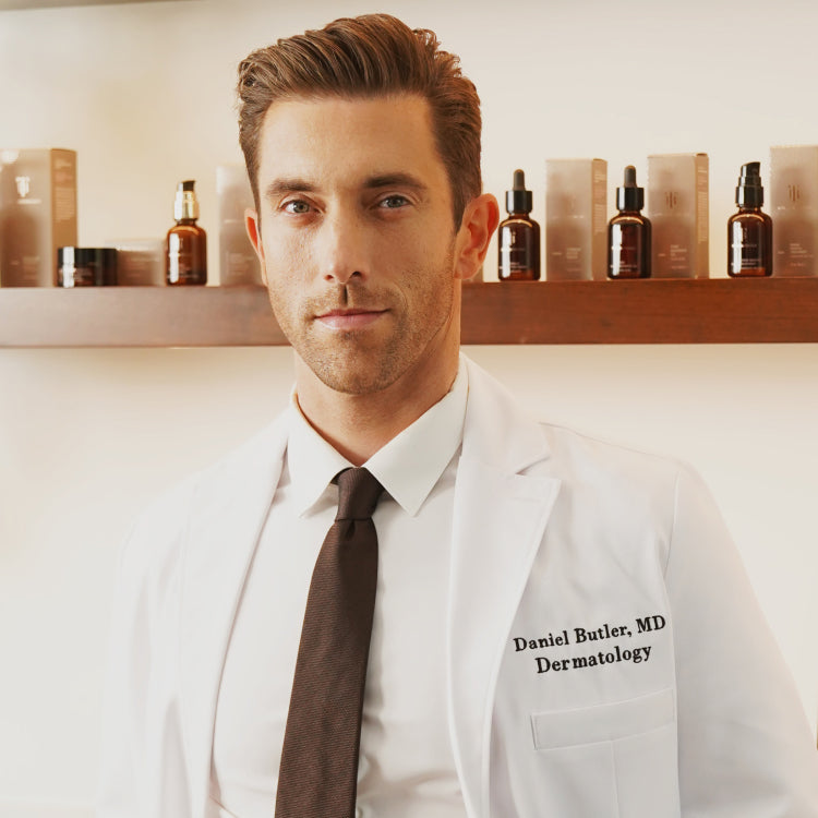 A man wearing a white lab coat embroidered with "Daniel Butler, MD, Dermatology" stands confidently in front of a wooden shelf. The shelf is lined with amber glass bottles and boxes of True Botanicals skincare products. He has neatly styled hair and a professional demeanor, with a neutral-toned background that emphasizes a clean, clinical setting.